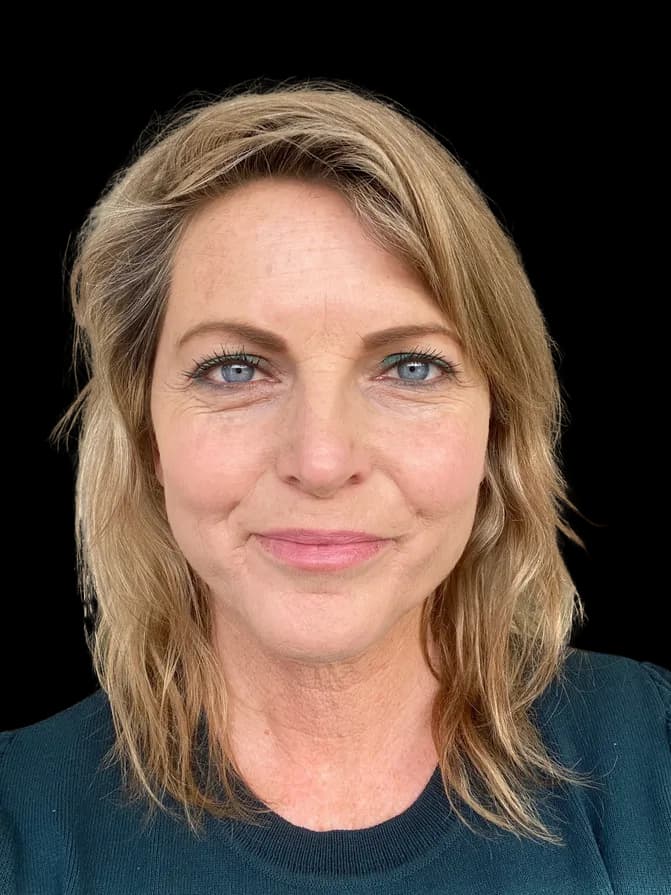 Middle-aged woman with blonde hair and blue eyes smiling slightly against a black background.